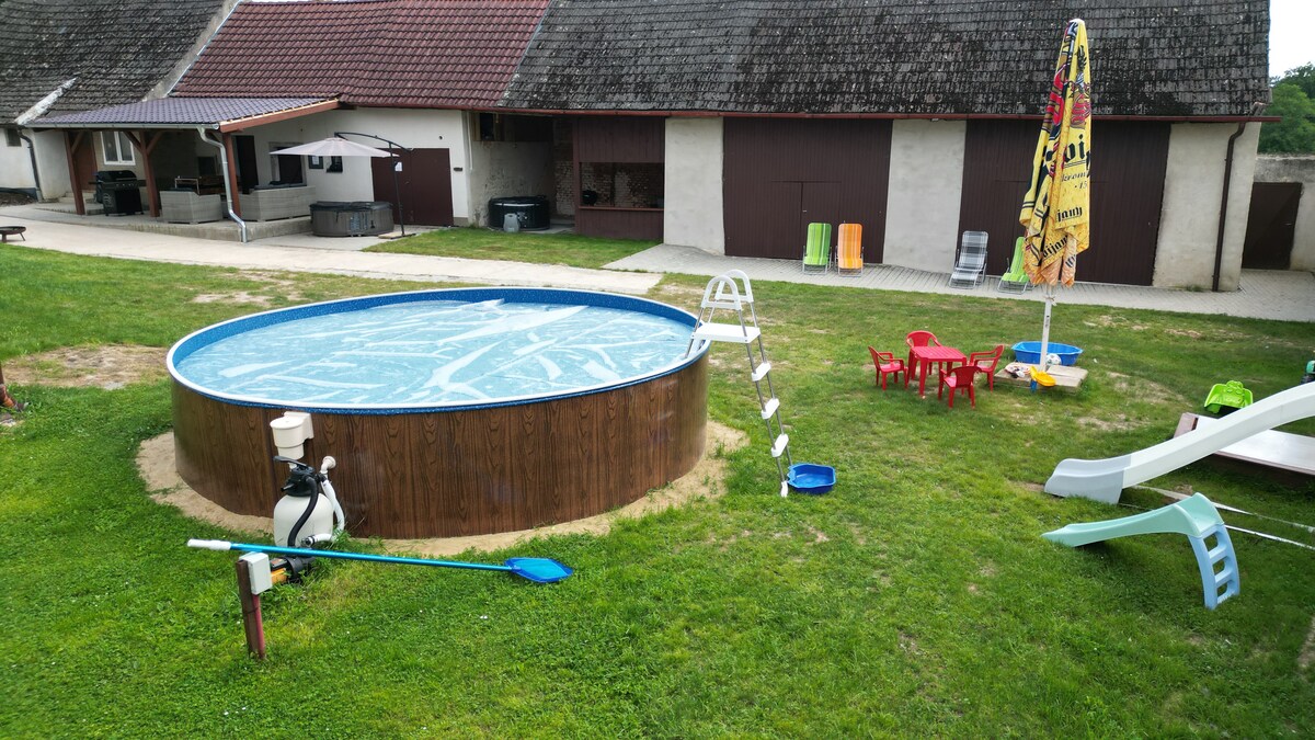 A round above-ground pool is situated in a grassy yard, surrounded by a wooden deck. Colorful plastic chairs and a slide are nearby, along with a yellow umbrella providing shade. The area is well-maintained, enhancing the peaceful outdoor setting.