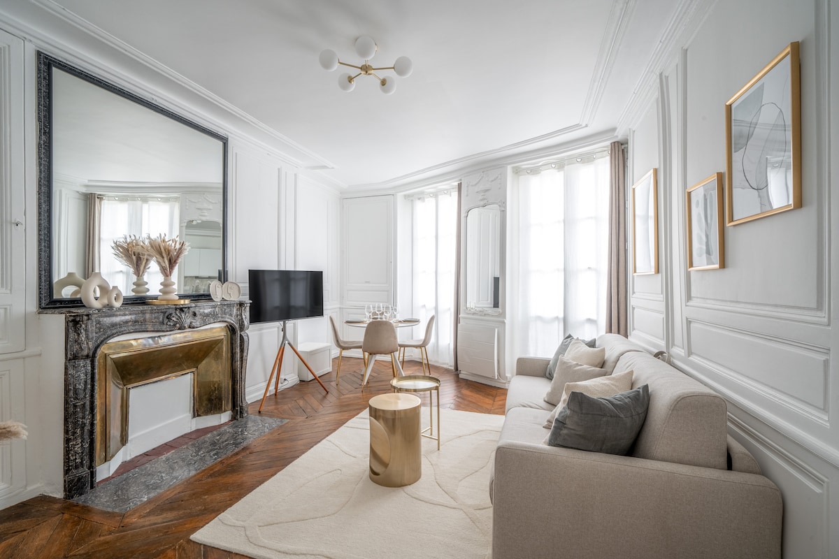 The living area features a neutral palette with elegant wall panels and large windows that allow natural light. A comfortable sofa is positioned beside a modern gold-accented coffee table. A television is mounted on the wall, and a decorative fireplace adds charm to the space.