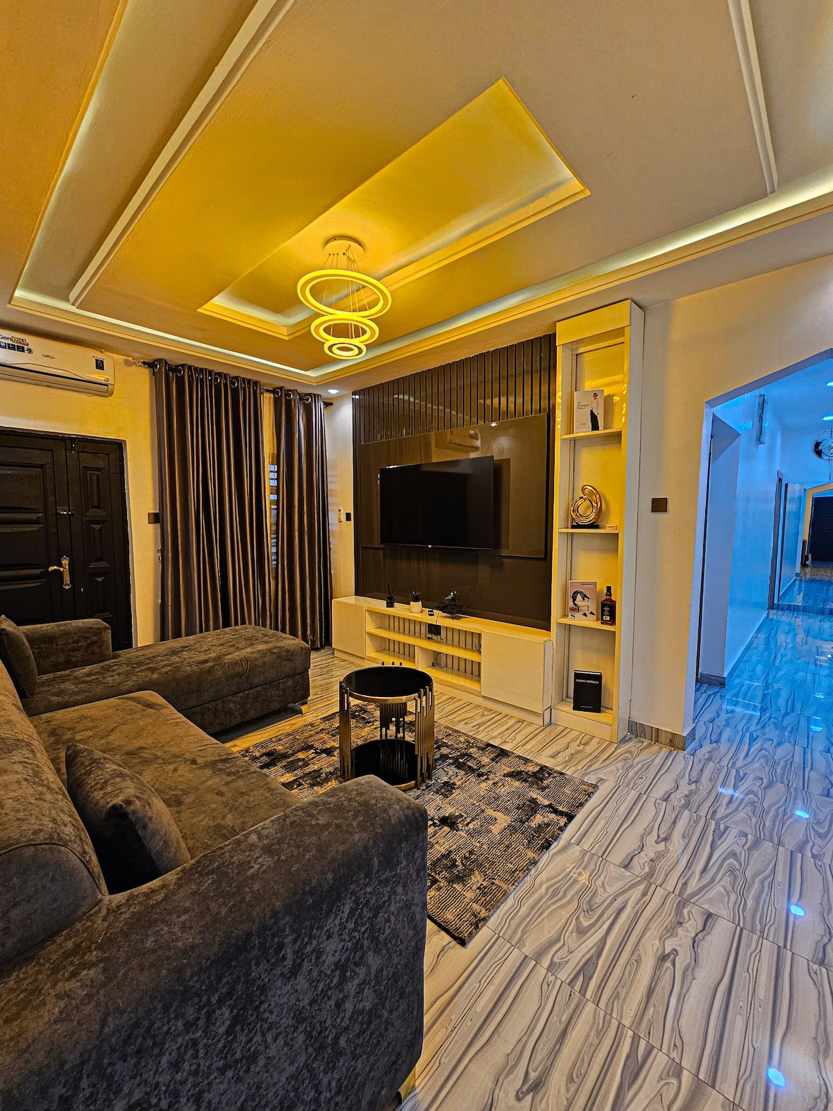 A stylish living area features a comfortable dark sofa and an inviting coffee table set on a patterned rug. A wall-mounted television is positioned above a built-in storage unit, while warm lighting from a modern ceiling fixture adds a soft glow to the space.