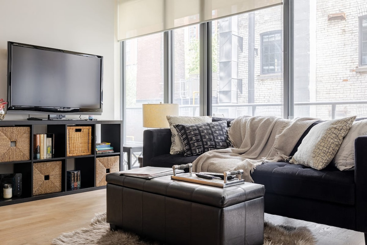A comfortable living area features a dark sofa adorned with a light blanket and decorative pillows. A wooden coffee table sits in front, accompanied by a storage ottoman. Large windows allow natural light to fill the space, highlighted by a television on a nearby console.