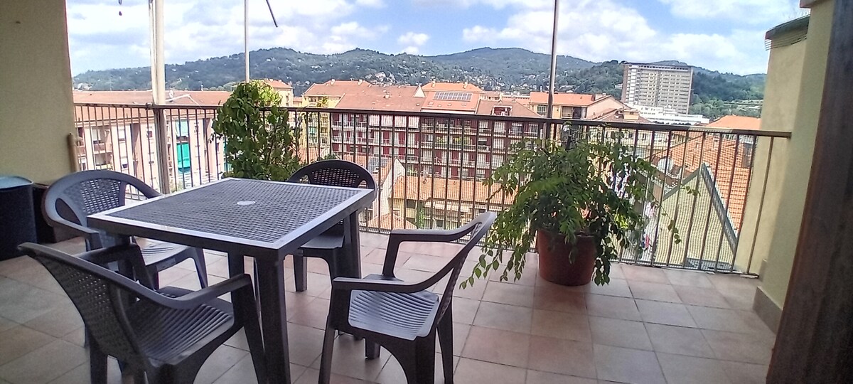 A private terrace features a square table surrounded by four chairs, offering views of the surrounding hills. Potted plants add greenery, while the vast skyline is visible in the background under a partly cloudy sky.