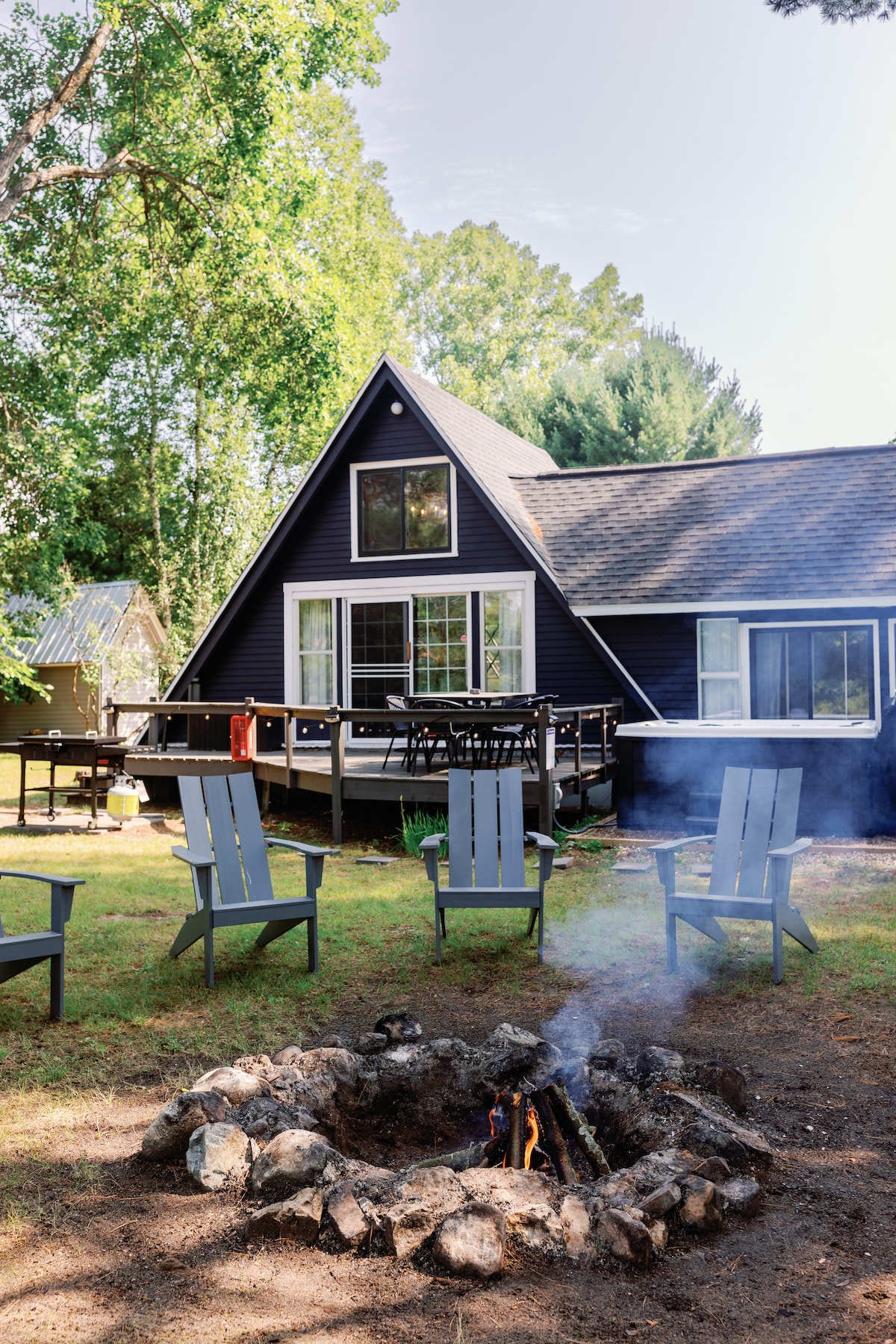 A charming A-frame cottage is set among tall trees, featuring large windows that offer a view of the outdoors. A circular stone fire pit is surrounded by several patio chairs, creating a perfect spot for gathering and enjoying warmth from the fire.
