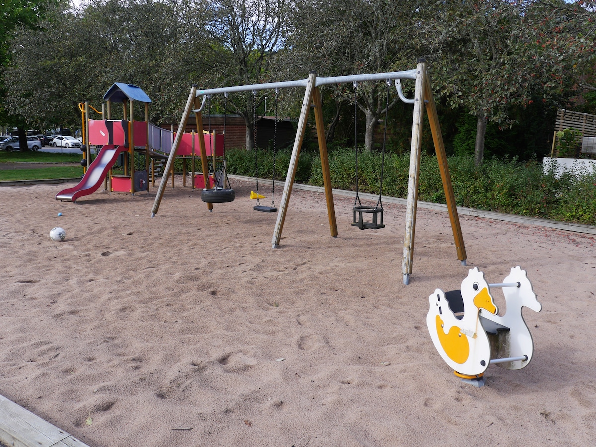 A communal playground is depicted, featuring swings set on sandy ground, along with a slide and a spring-mounted ride shaped like ducks. Lush green trees and open space provide a natural backdrop for children to enjoy outdoor activities.