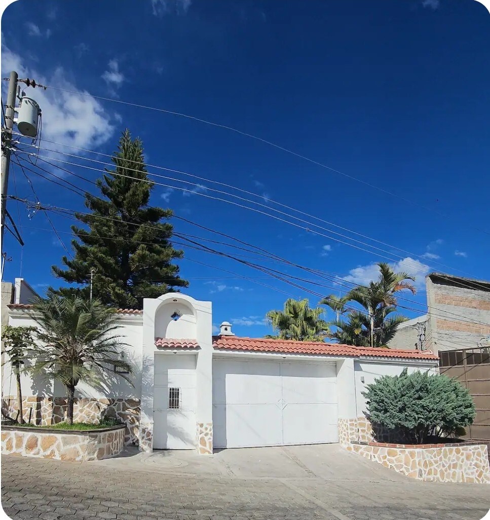 The exterior of a white, single-story house is presented, featuring a red-tiled roof and a large garage door. Lush green palm trees and shrubs surround the entrance, with a clear blue sky overhead, adding to the property’s welcoming appearance.