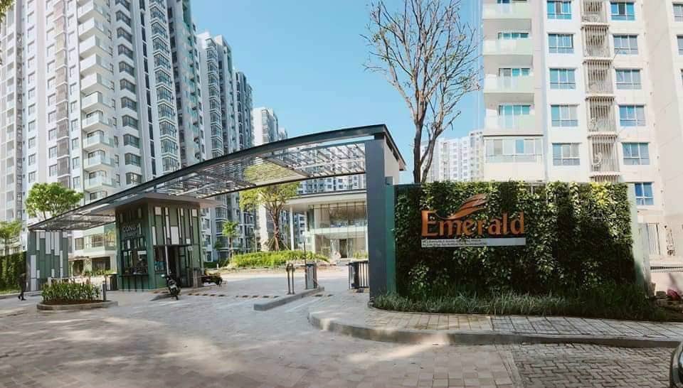 An entrance is featured with a modern design, showcasing a glass canopy supported by sleek metal frames. The exterior wall is adorned with greenery, while the name 'Emerald' is prominently displayed. Surrounding high-rise buildings and a clear blue sky complete the scene.