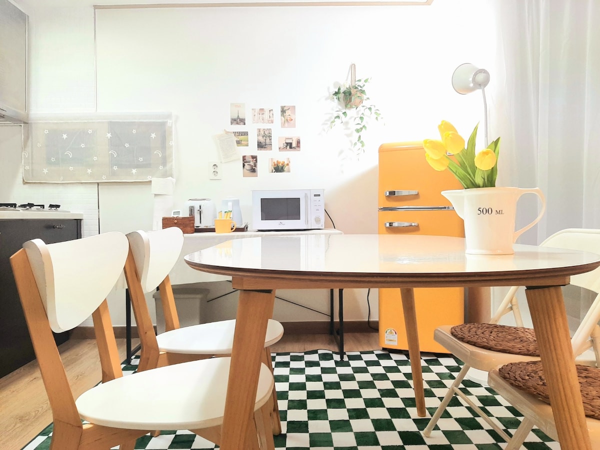 A dining area features a light wood table surrounded by four white chairs. A vintage-style orange refrigerator is visible in the background, along with a microwave. Fresh yellow tulips are displayed in a pitcher at the center of the table, enhancing the inviting atmosphere.