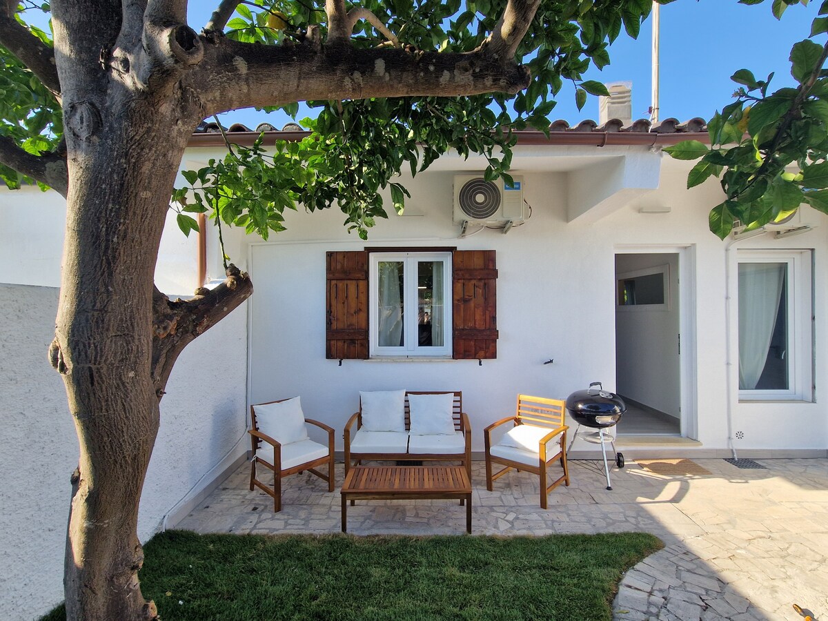 A shaded outdoor seating area is presented with a wooden coffee table flanked by two white chairs and a bench. The space is adjoining a white wall and features a BBQ grill on one side, with lush greenery enhancing the setting.