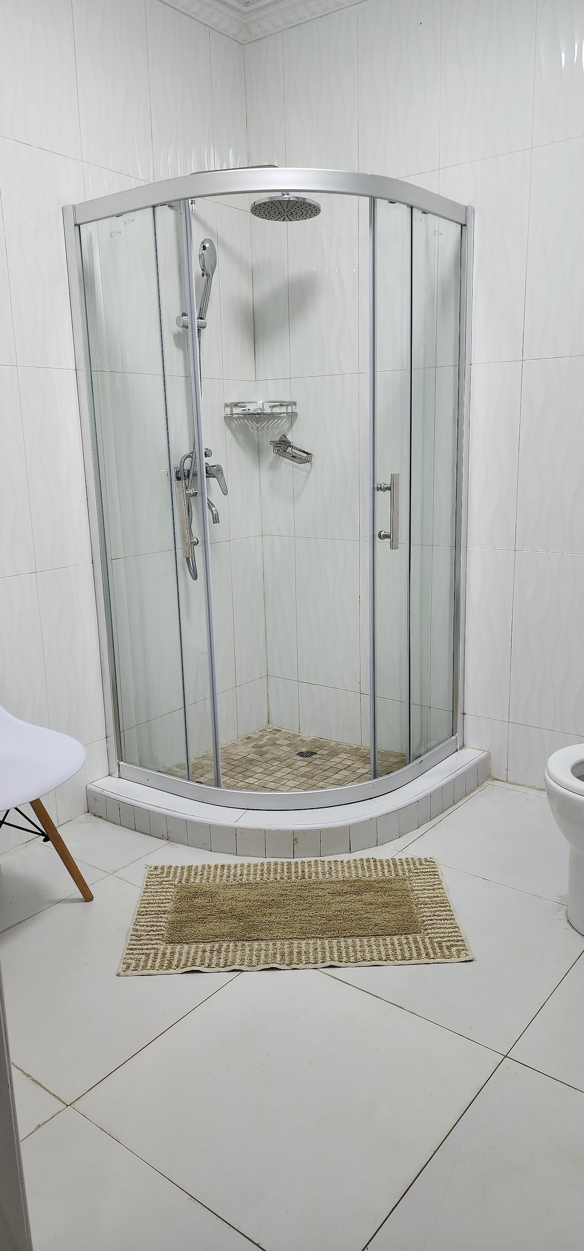 A modern shower enclosure is featured, surrounded by white tiled walls and a light-colored tiled floor. A showerhead and handheld fixture are visible within the glass doors, while a woven mat is placed at the entrance. A toilet is positioned nearby.