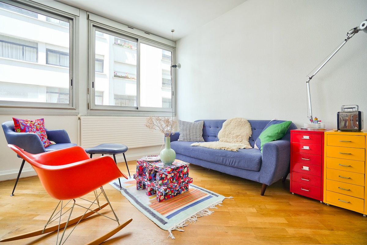A bright living area features a cozy blue sofa and a red rocking chair, complemented by a colorful patterned coffee table. Large windows allow natural light to fill the space, while decorative elements, including a plant and vibrant cushions, enhance the welcoming environment.