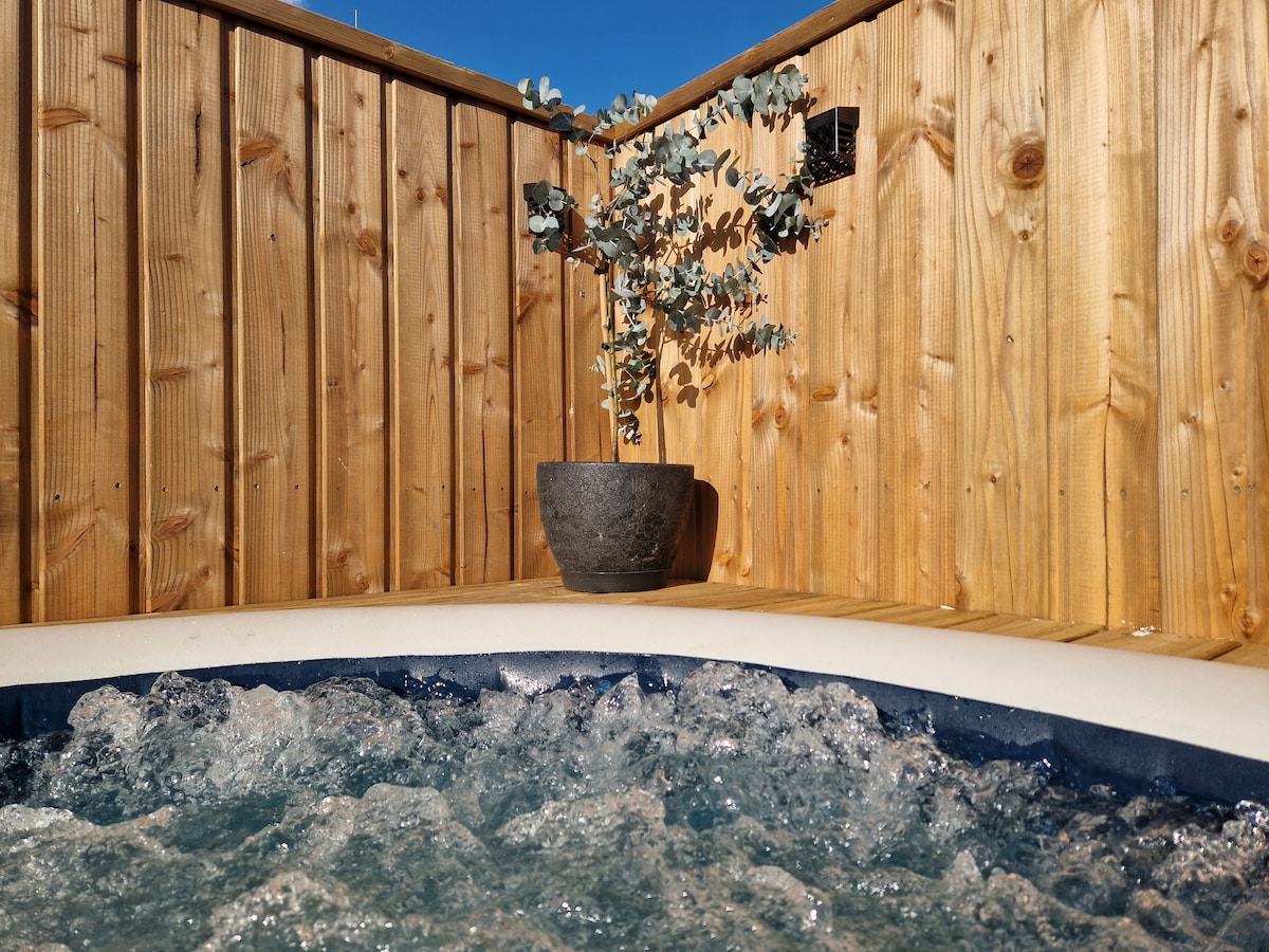 A corner of the outdoor spa area is depicted, featuring a bubbling spa surrounded by wooden fencing. A potted plant is positioned against the fence, adding a touch of greenery to the serene setting.