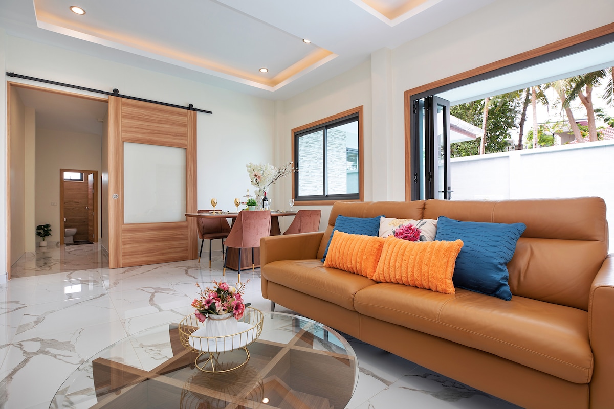 A modern living area features a sleek leather sofa adorned with colorful cushions. A glass coffee table sits in front, while large windows allow natural light to illuminate the space. A dining area is visible in the background, showcasing a table set for four.