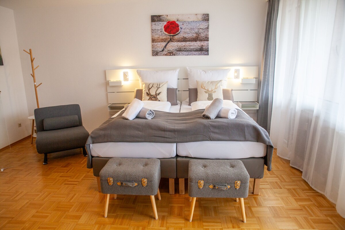 A spacious sleep area features two elevated beds with neutral bedding and decorative throw pillows. A modern artwork adorns the wall above. Two small stools are placed at the foot of the beds. A cozy chair is situated in the corner, alongside natural light streaming through the sheer window curtains.