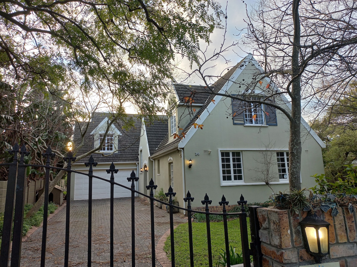A charming two-story home with a pitched roof is visible, surrounded by greenery. The front yard features well-kept grass and a gated entrance. A garage is located on the left, and a tree provides shade to the area, creating a welcoming exterior.