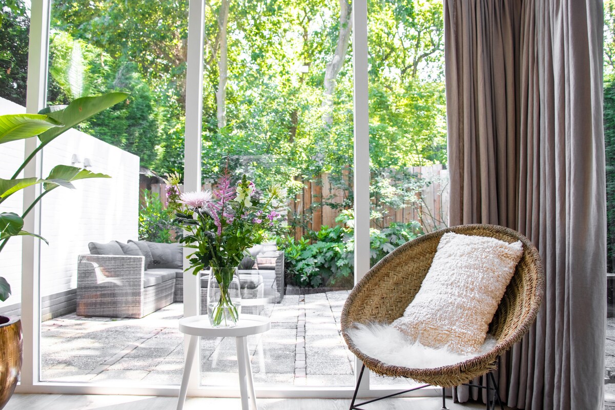 A bright seating area features a woven chair and a small table with a vase of flowers. Large glass doors provide a view of a lush garden, while soft curtains frame the natural light streaming into the space.