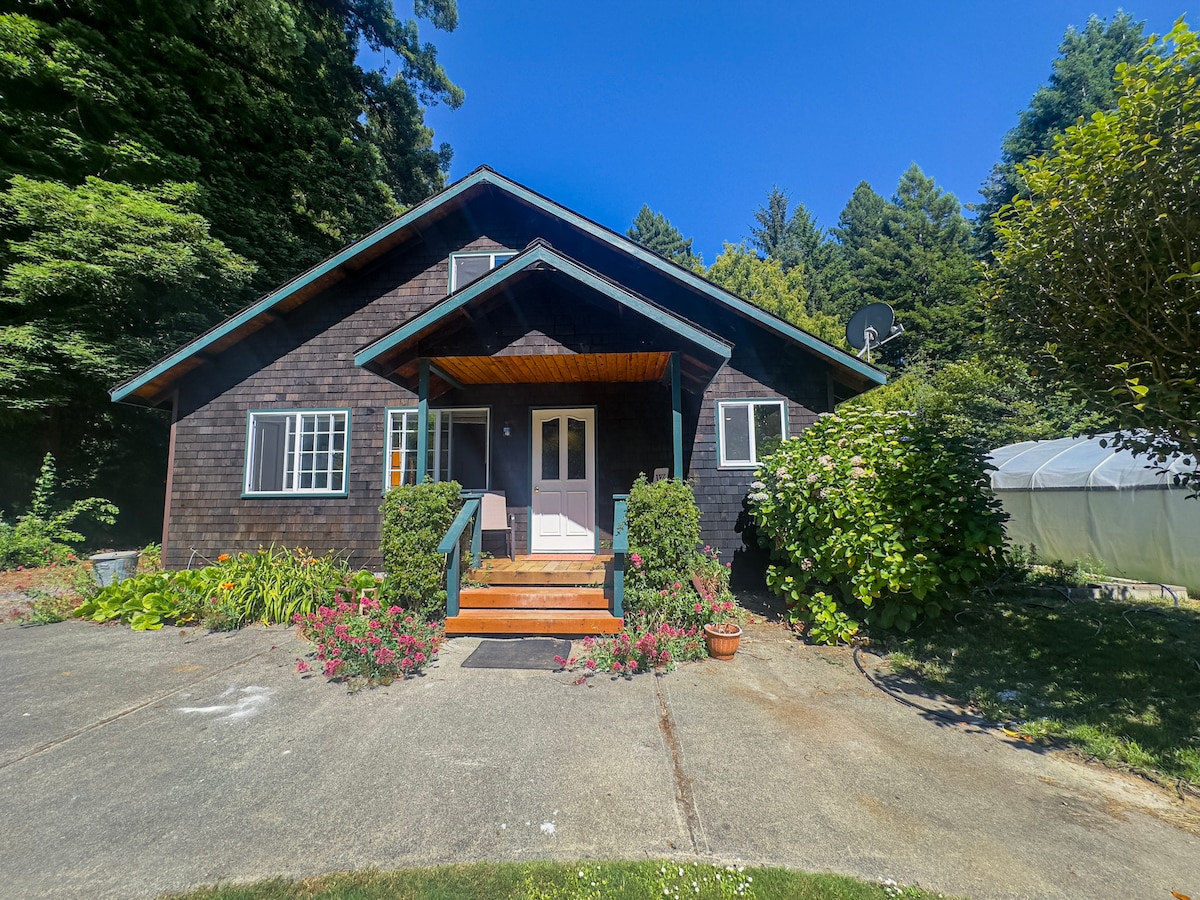 The exterior of the cottage is set against lush greenery, featuring a dark wood facade and a welcoming front porch. A pathway leads to the entrance, bordered by vibrant garden flowers, while trees rise in the background, completing the serene setting.