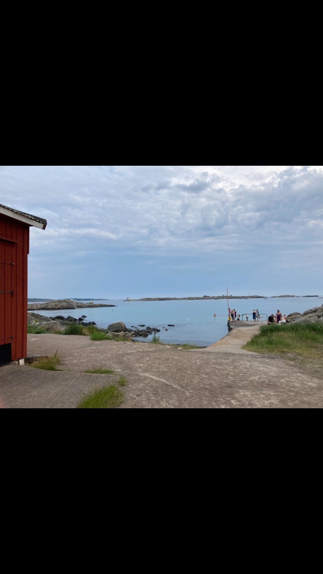 A red wooden structure is visible at the edge of a rocky shore, leading to calm waters. Groups of people are seen enjoying the scenery, with distant islands and a lighthouse appearing along the horizon under a cloudy sky.