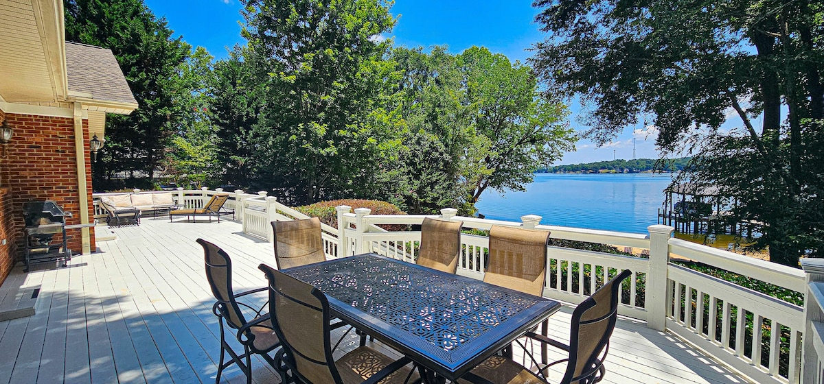 An outdoor deck space features a large, dark metal dining table surrounded by six brown chairs. Lush greenery and a serene lake view provide a tranquil backdrop. Sun loungers are positioned nearby, inviting relaxation in the sun-dappled setting.