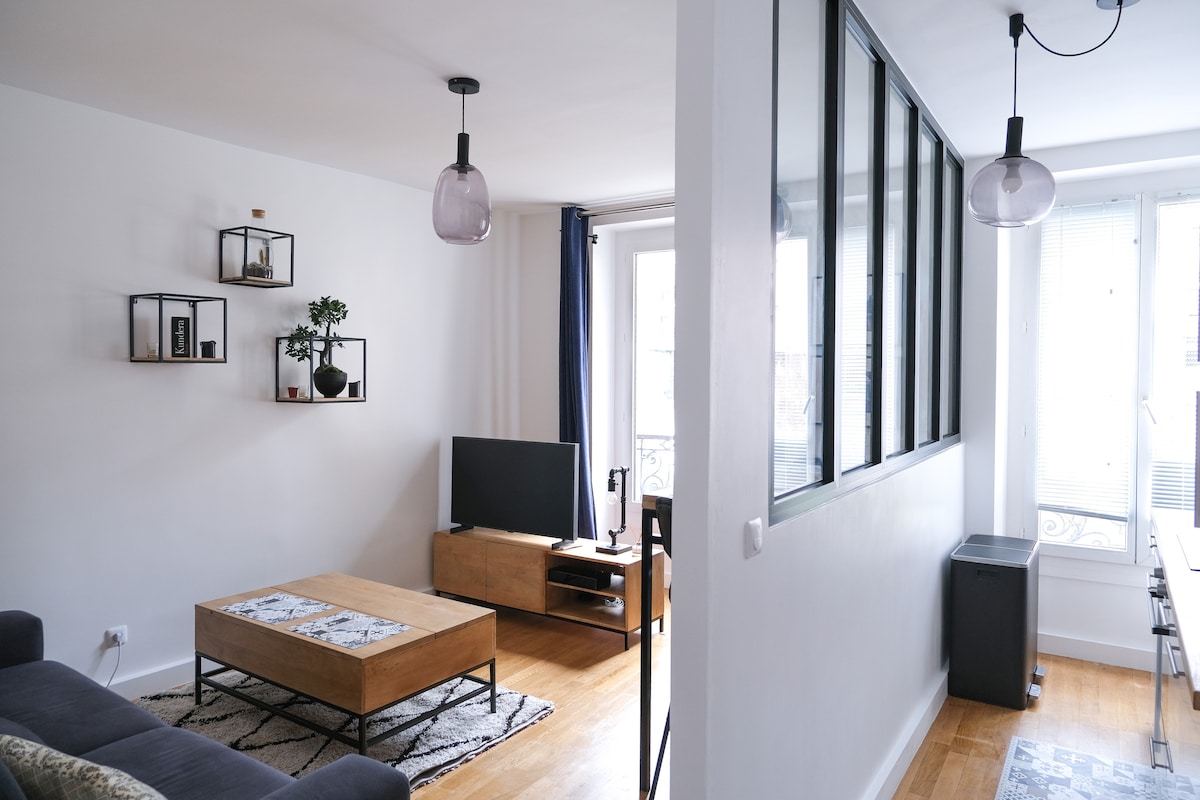 The living area features a low wooden coffee table with a decorative runner, a sleek modern TV stand, and a television. Stylish shelves adorn the wall, holding various decor items. Natural light floods in through large windows, contributing to a bright atmosphere.