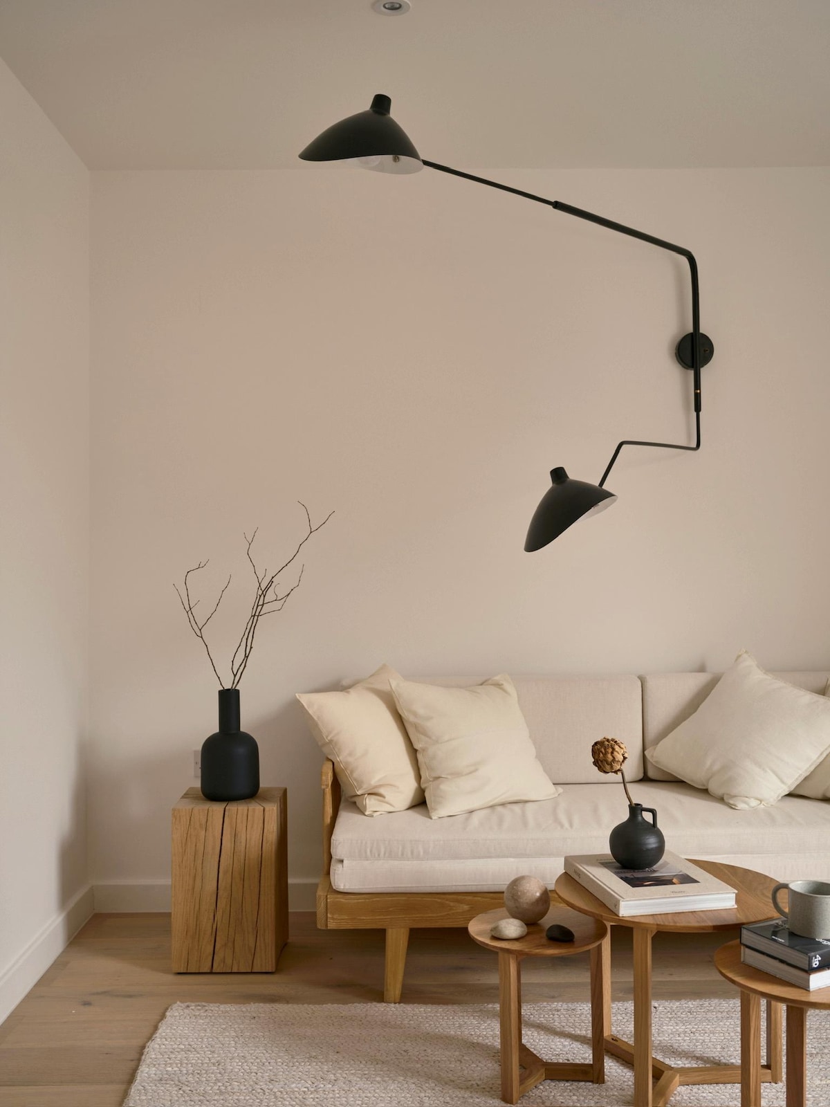 A soft minimal living area is shown, featuring a light beige couch adorned with plush cushions. A minimalist wooden side table holds decorative objects, while a black floor lamp arcs overhead, illuminating the space. A slender vase with branches adds a gentle touch to the decor.