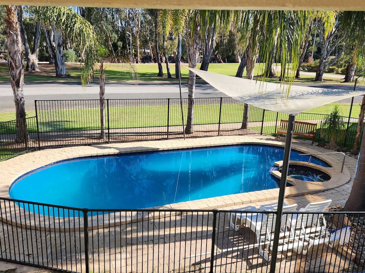 A refreshing outdoor swimming pool is framed by a spacious patio area. Surrounding palm trees provide shade, while a shaded lounge area with reclining chairs is positioned nearby. A clear view of the manicured lawn beyond the pool is also visible.