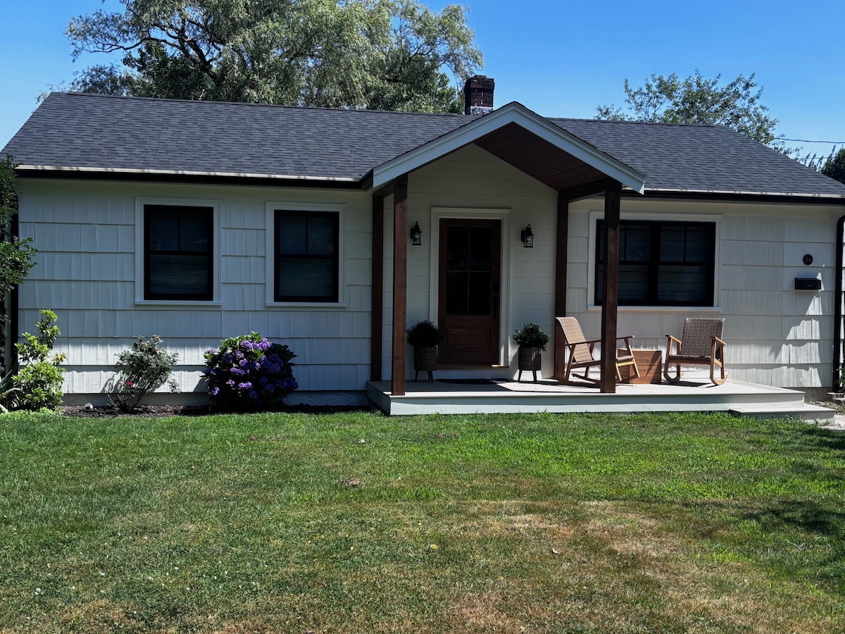 The charming cottage exterior features a welcoming porch with two rocking chairs and lush greenery surrounding it. The light-colored facade is complemented by dark windows and a pitched roof, creating a cozy, inviting appearance. A manicured lawn enhances the overall setting.