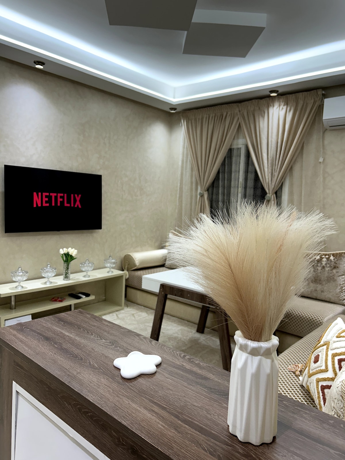 A modern living room features a wall-mounted TV displaying the Netflix logo. Elegant curtains frame a large window, while decorative items, including a vase with dried floral accents, enhance the space. A comfortable seating area is positioned around a neutral-colored coffee table.