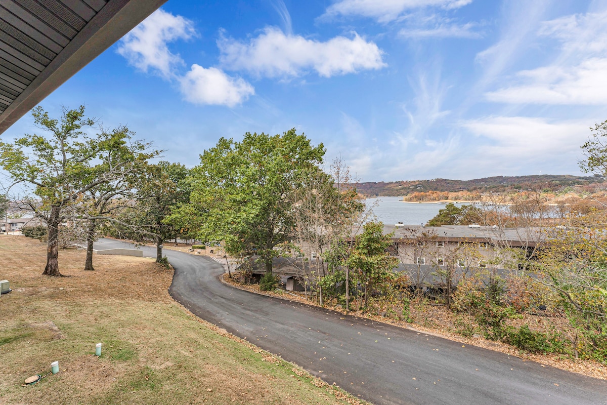 A scenic view is displayed from the balcony, featuring a winding road lined with trees leading towards Table Rock Lake. The clear blue sky and gentle clouds complement the landscape, while the nearby resort buildings are nestled among the greenery.