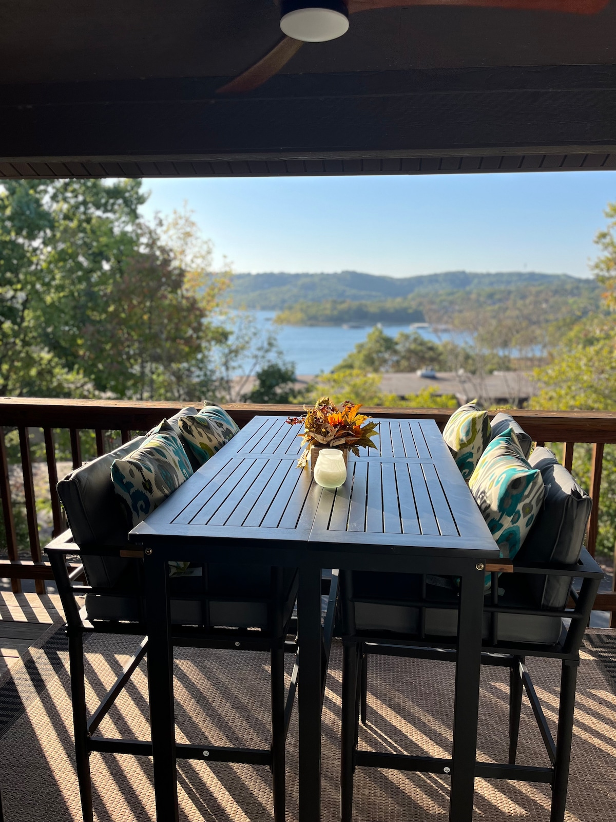 A dining table with cushioned chairs is positioned on a private balcony, offering a view of the serene lake and surrounding trees. A small vase with seasonal flowers adorns the table, inviting leisurely meals in a tranquil outdoor setting.