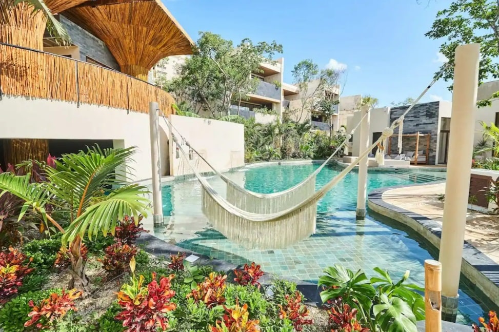 A serene pool area is surrounded by lush tropical landscaping, featuring vibrant plants and a comfortable hammock suspended over the water. Natural materials are used in the nearby structures, harmonizing with the relaxing ambiance of the outdoor space.