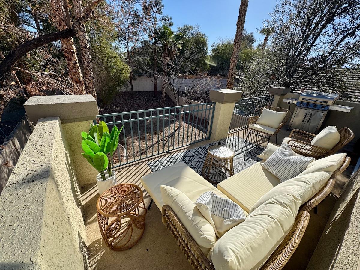 A spacious patio is furnished with a cushioned seating arrangement and a small round table. A BBQ grill is positioned in the corner, surrounded by greenery and a view of the serene backyard, creating an inviting outdoor space for relaxation.