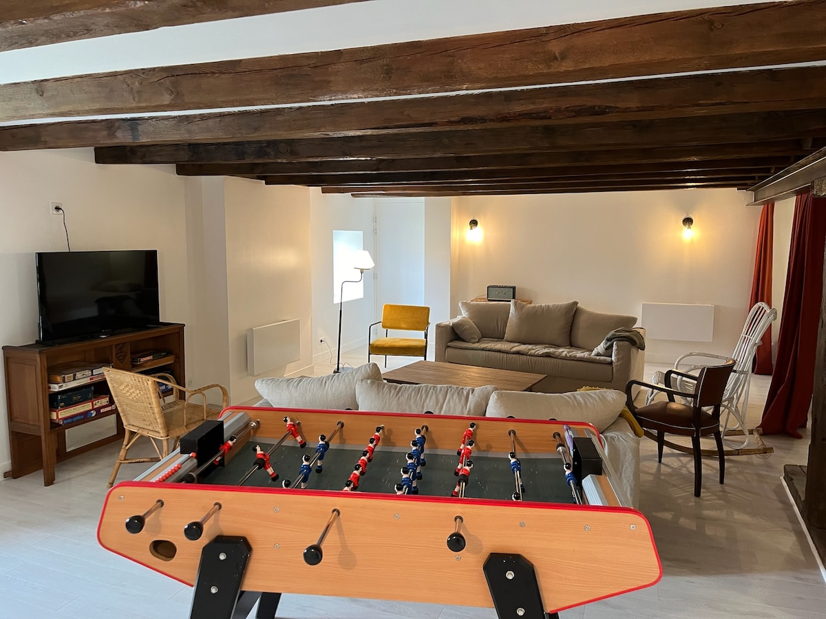 A spacious common area features wooden beams above, with a foosball table in the foreground. The seating includes a soft sofa, armchair, and rocking chair, while a TV and bookshelf are visible, creating an inviting space for relaxation and entertainment.