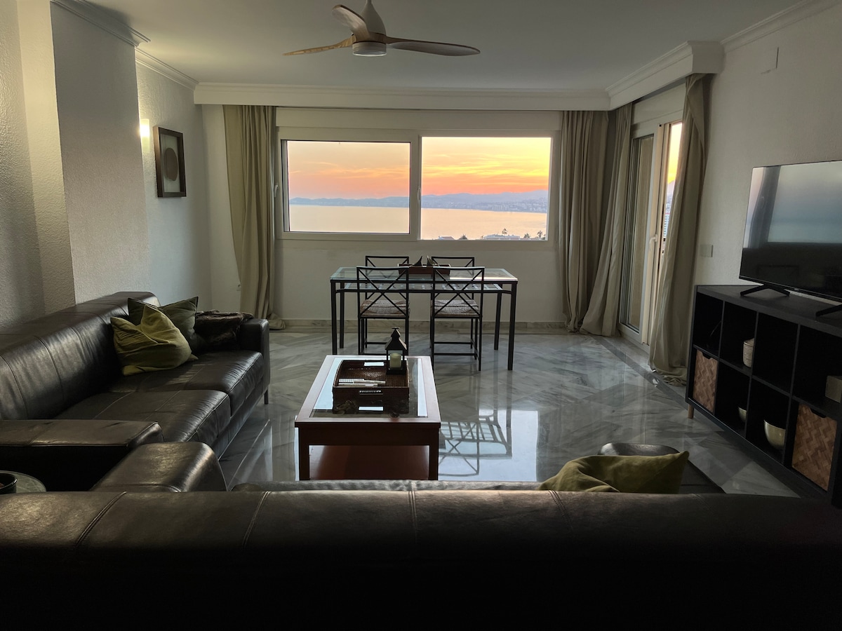 A spacious living area features two large sofas arranged around a coffee table, with a dining table positioned by the window. Soft curtains frame the expansive windows, allowing views of the sunset over the water. A television is mounted on the wall, enhancing the modern ambiance.
