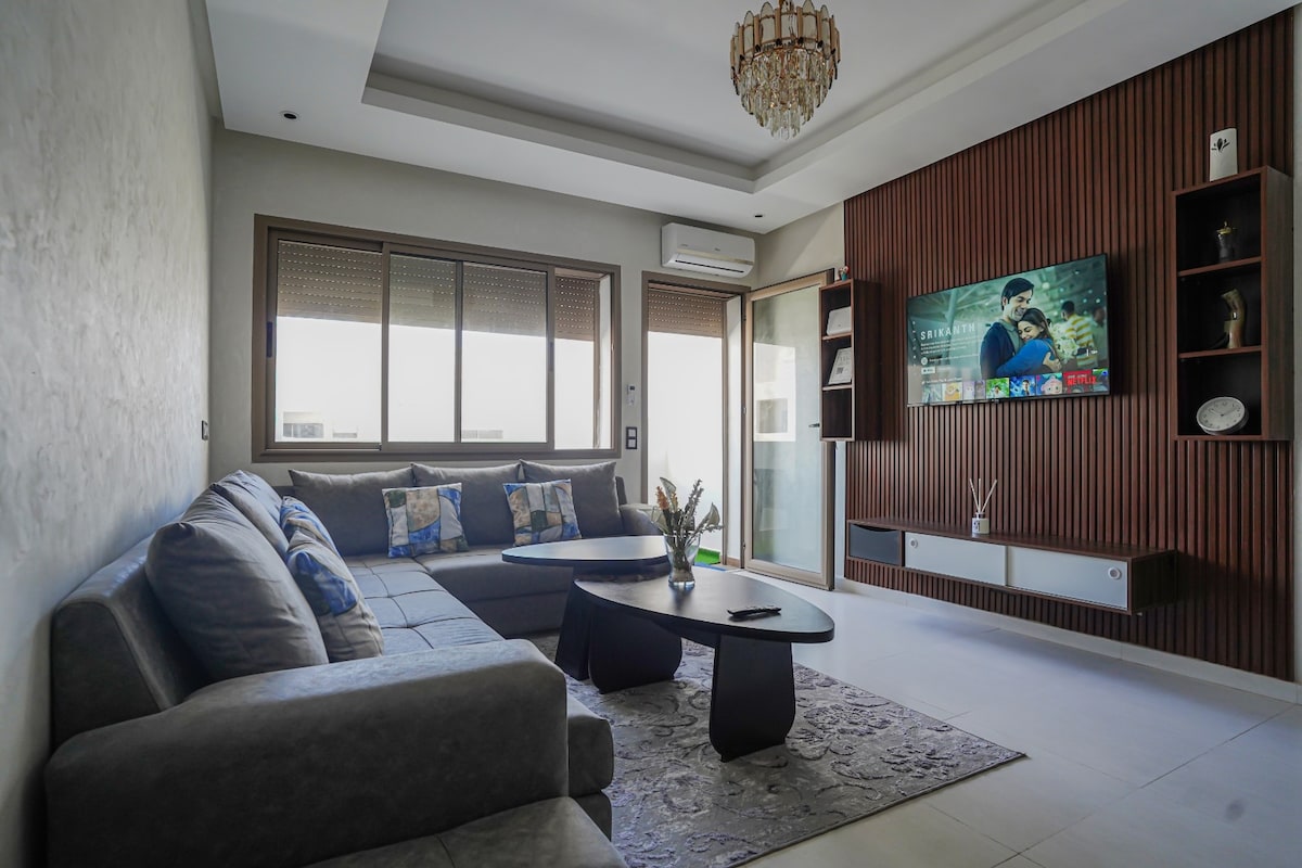 The living area is furnished with a large, comfortable sectional sofa and a modern coffee table. A flat-screen TV is mounted on the wall, displaying various content. Natural light filters through the windows, enhancing the space's brightness and openness.