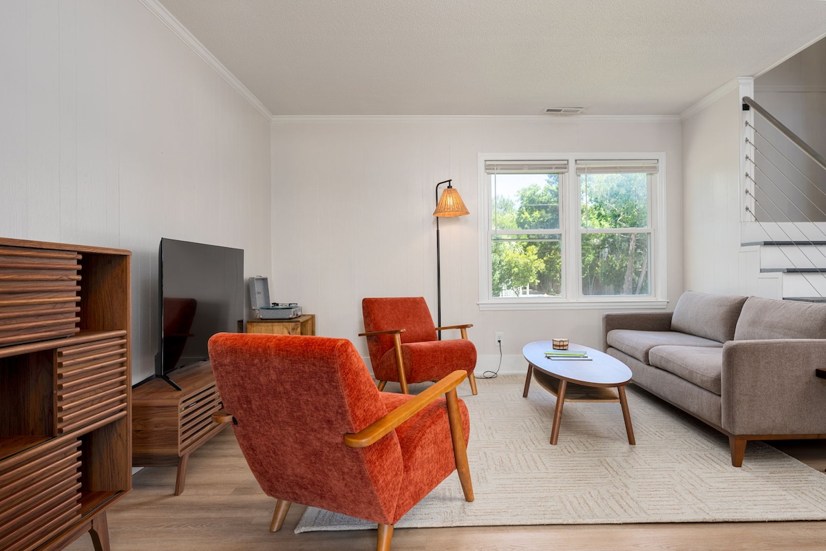 The living area features a modern design with a light-colored rug under a round coffee table. Two orange armchairs provide comfortable seating next to a gray sofa. A television is positioned on a wooden media console, with large windows allowing natural light to enter the space.