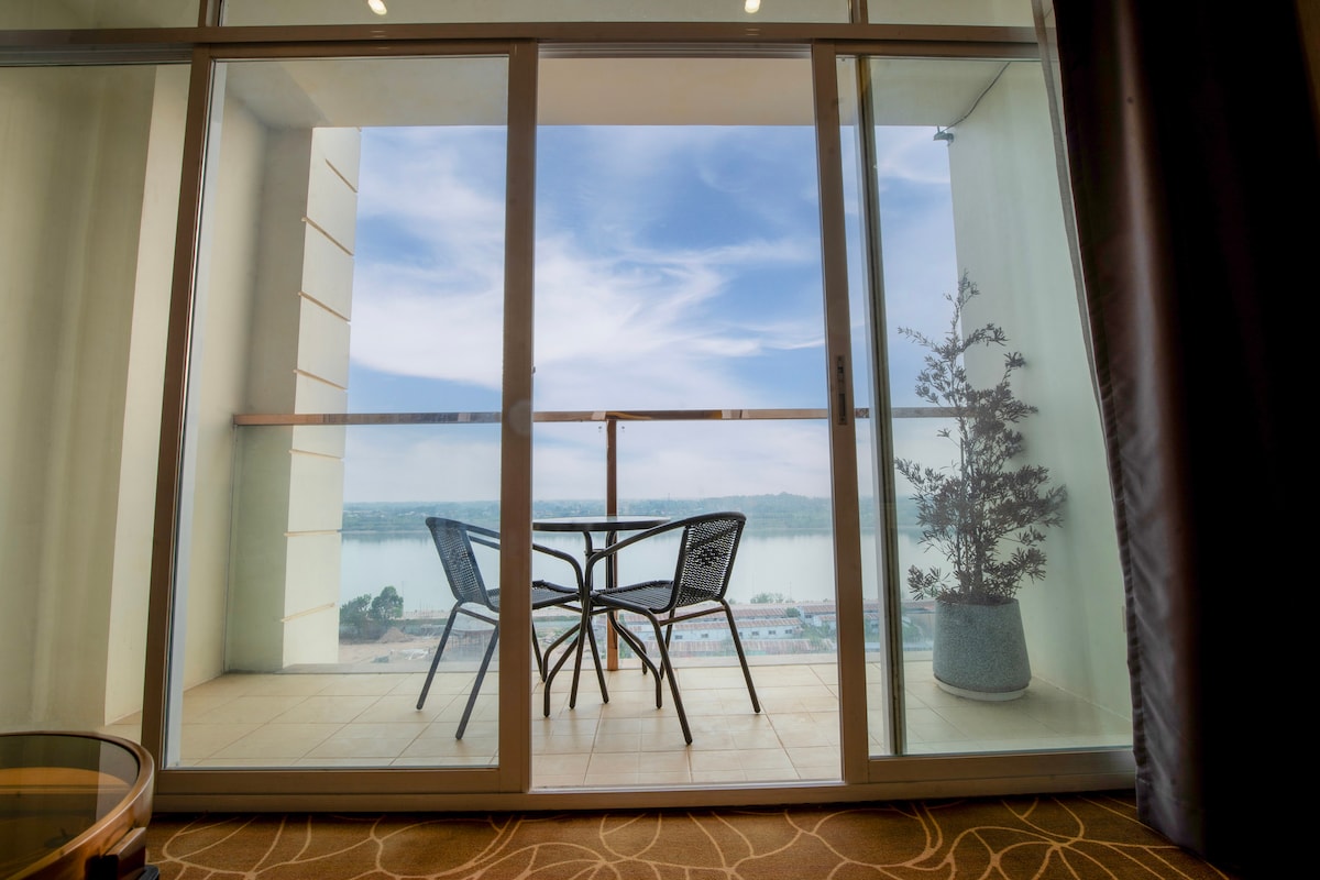 A private balcony is visible, featuring a small round table surrounded by two chairs. The expansive glass doors frame a scenic view of the landscape, with light filtering through while a potted plant adds a touch of greenery.