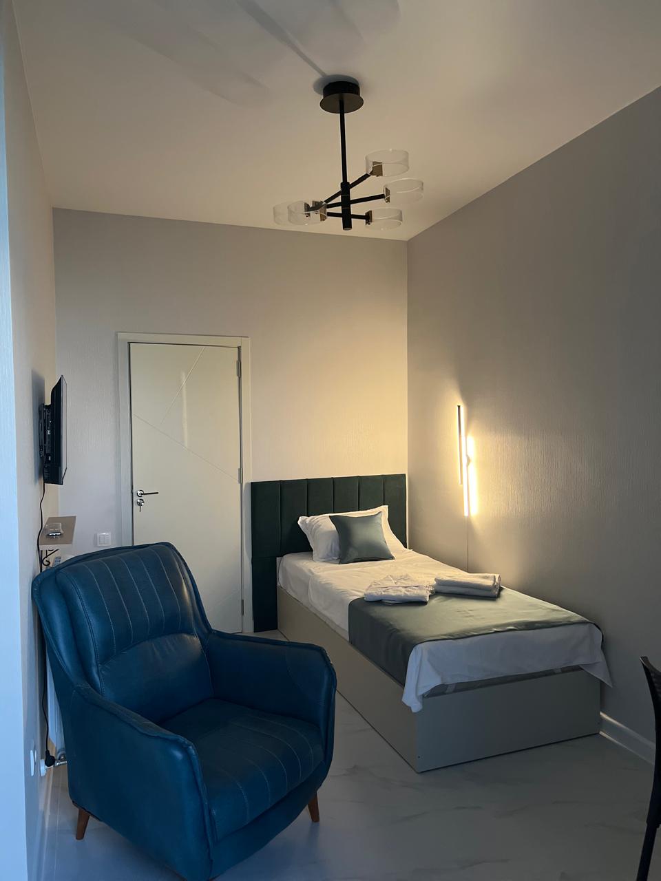A bright room features a single bed with a light blue throw and matching linens. A comfortable armchair in deep teal is positioned beside the bed. A modern light fixture hangs from the ceiling, while a TV is mounted on the wall opposite. Natural light is subtly filtered through the room.