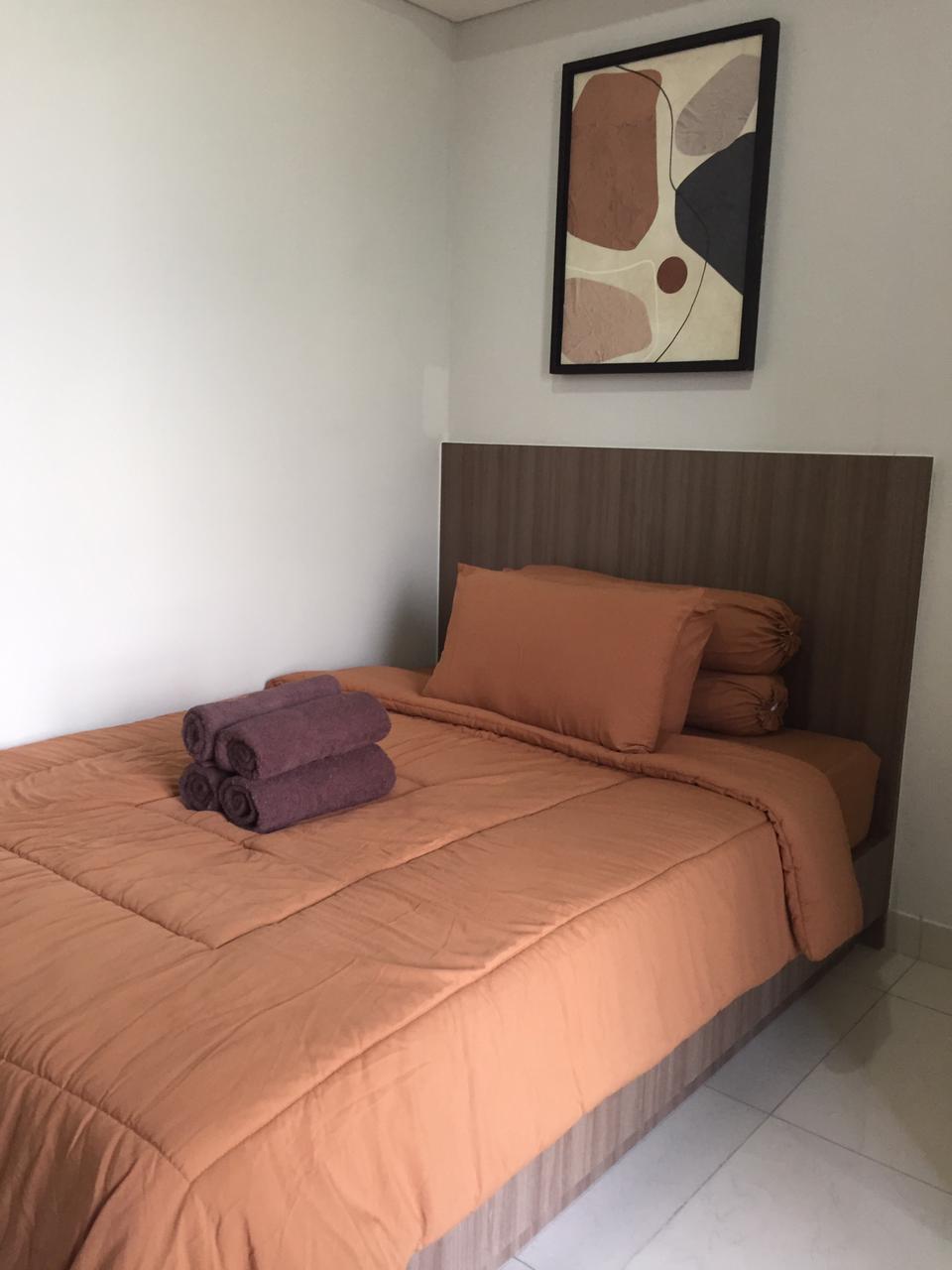 A spacious bed dressed in warm orange bedding is positioned against a sleek wooden headboard. Neatly rolled towels in deep purple are placed on the bed, complementing the modern wall art above. Natural light highlights the clean lines and minimalist decor.