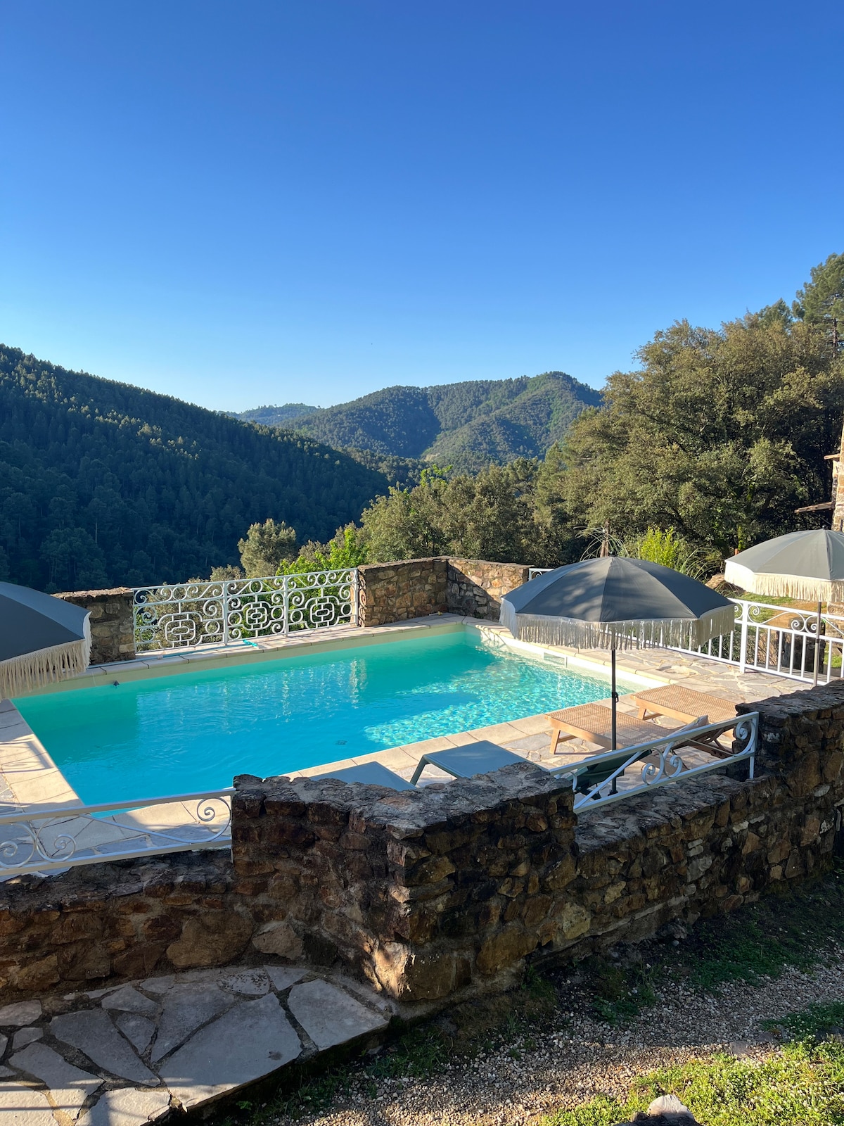 A serene swimming pool is surrounded by stone walls and lush green hills. Sun loungers are positioned under gray umbrellas, offering shade. The clear blue sky reflects on the water, creating a tranquil ambiance. The inviting pool area overlooks the stunning mountainous landscape.