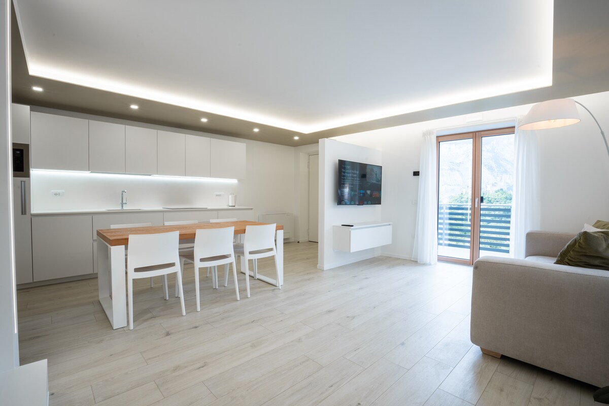 A modern and spacious living area features a sleek kitchen with white cabinetry and a wooden dining table accompanied by four white chairs. Natural light floods the space through glass doors leading to a balcony, while a comfortable sofa and a wall-mounted TV enhance the atmosphere.