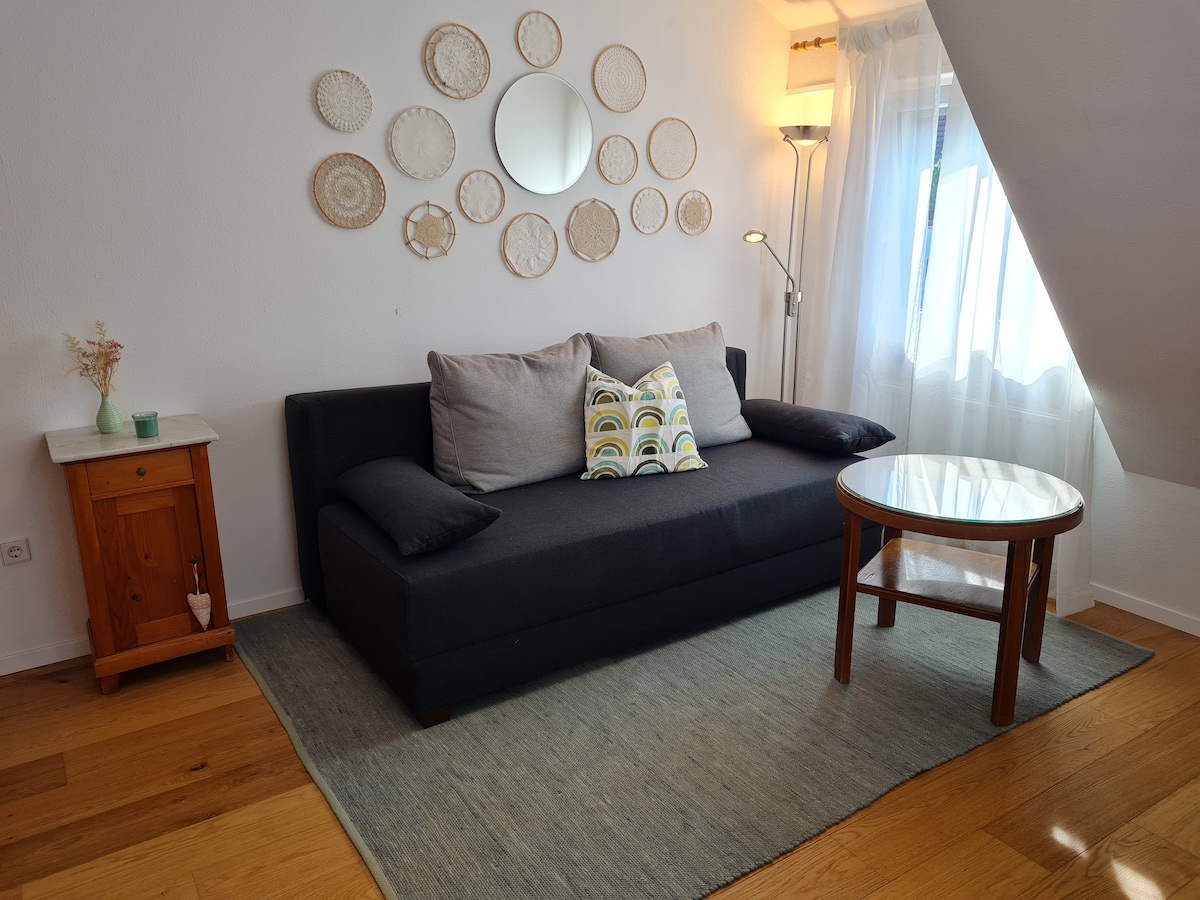 A cozy living area features a dark sofa with decorative cushions, paired with a round wooden table. A textured rug anchors the space. Wall decor includes circular woven pieces and a round mirror, while natural light filters through a sheer curtain beside a window.