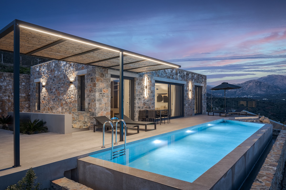 An outdoor area showcases a private pool set against a serene mountain backdrop, with lounge chairs positioned alongside. The villa's modern design includes stone walls and recessed lighting, creating an inviting atmosphere as the sun sets, illuminating the surroundings.