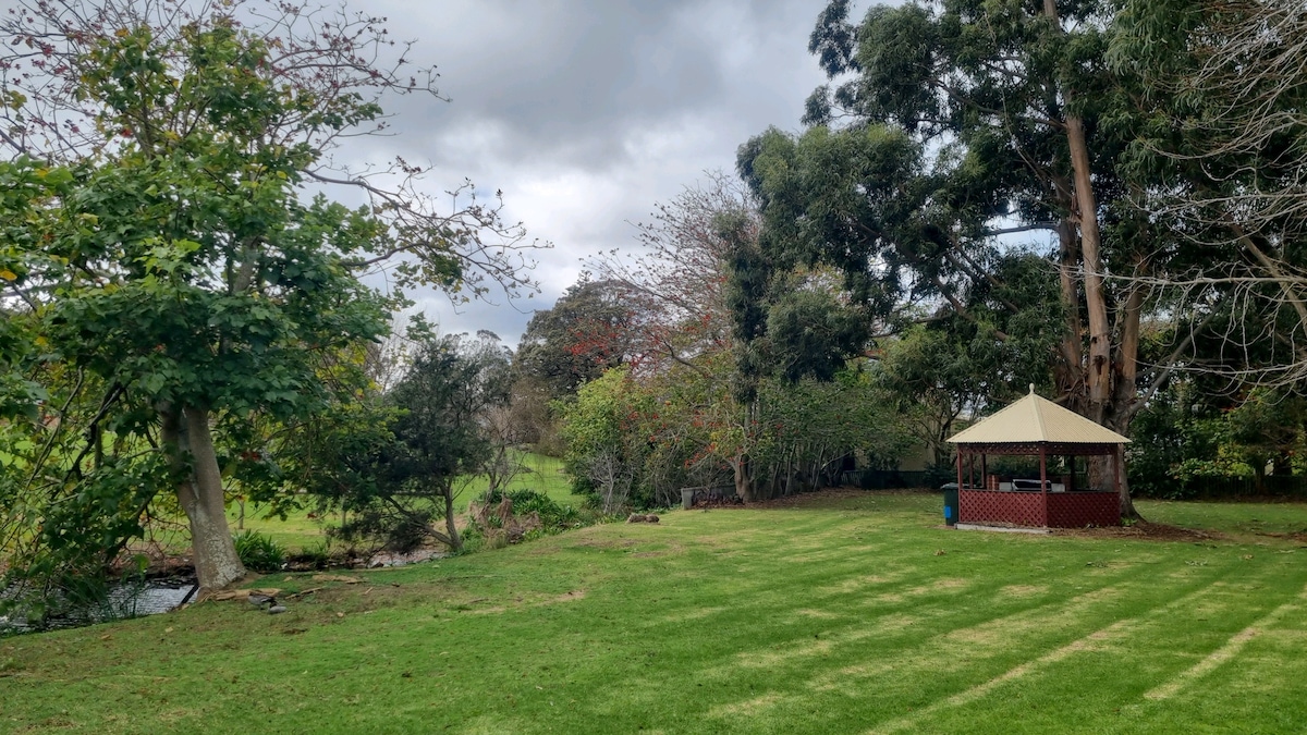 A spacious lawn area is framed by trees, providing a tranquil setting. A gazebo is situated on the right, accompanied by lush greenery. The landscape includes a small pond, and the sky is overcast, adding a serene atmosphere to the space.