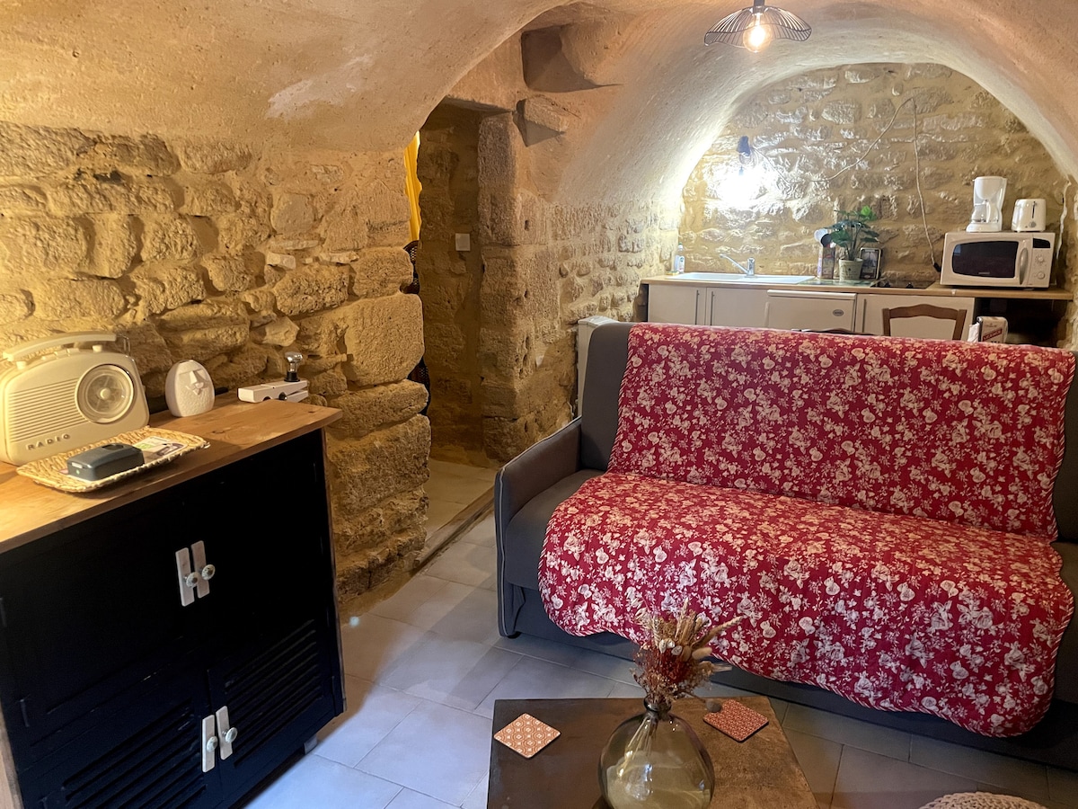 A cozy living area features a floral-patterned sofa bed, complemented by a wooden side table and vintage-style radio. Stone walls contribute to the character of the space, with a kitchenette visible in the background, showcasing modern appliances against rustic elements.