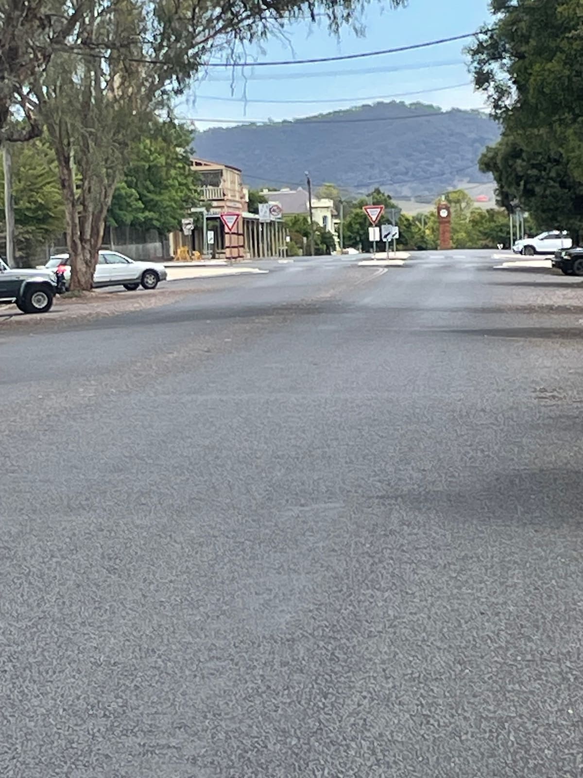 A quiet, tree-lined street is depicted, showcasing wide open spaces and several parked vehicles. The backdrop features distant hills under a clear sky, contributing to the serene atmosphere of the area. Street signs and lampposts are visible, emphasizing the relaxed pace of the neighborhood.