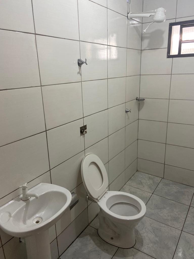 The bathroom features a tiled interior with a showerhead, a toilet, and a sink. Natural light enters through a small window, illuminating the clean, bright space. The design is simple and functional, offering essential amenities for shared use.
