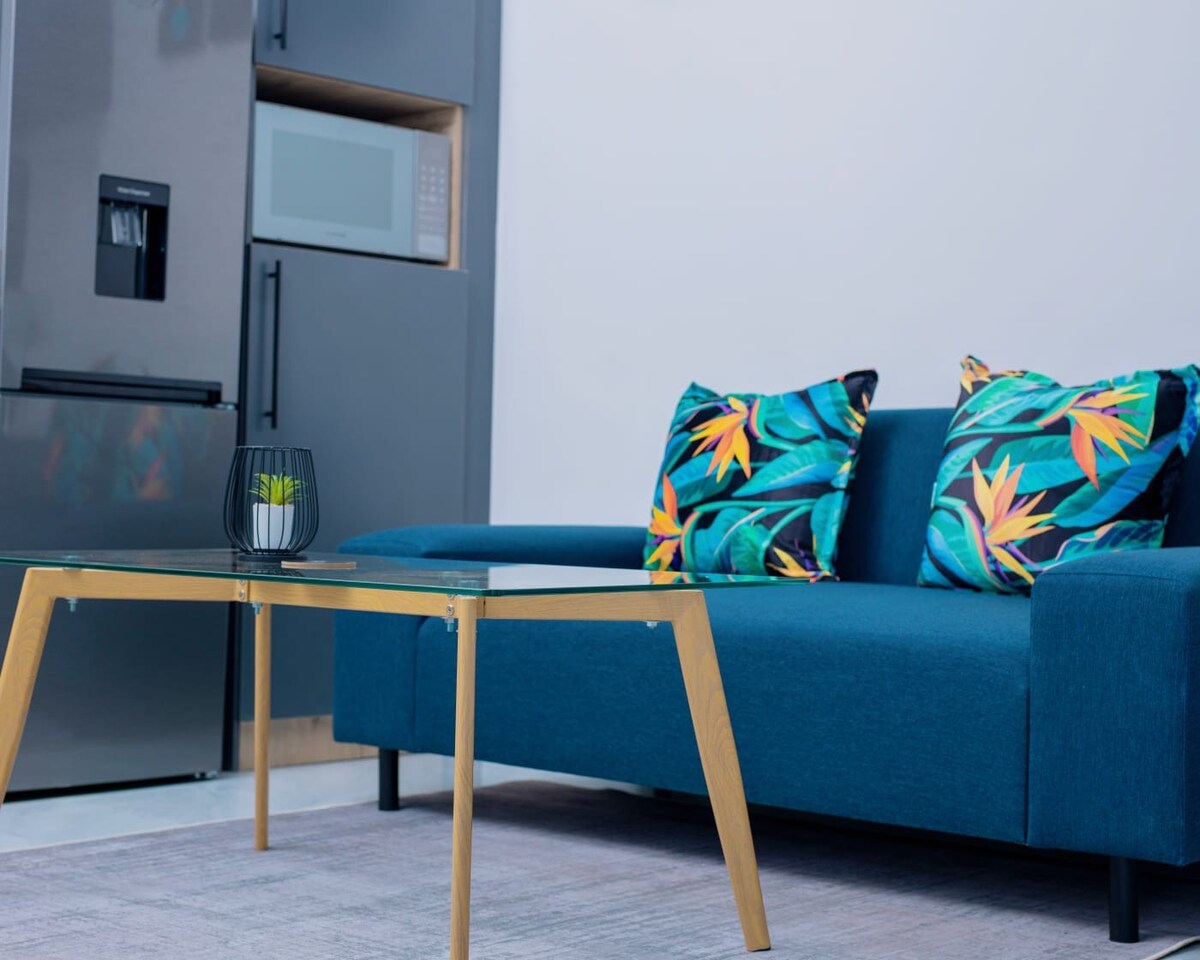 A modern living area features a stylish blue sofa adorned with tropical-patterned cushions. A glass coffee table with golden legs sits in front of the sofa, accompanied by a small decorative plant. The background displays a sleek refrigerator and a microwave.