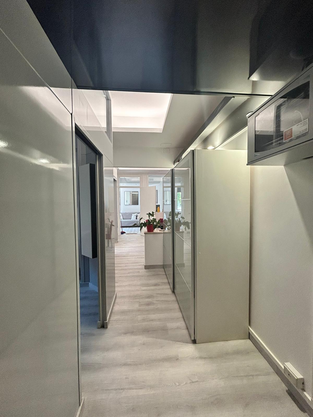 An open hallway is presented, featuring sleek, shiny walls and an illuminated ceiling. Glass doors on one side allow visibility into the adjacent rooms. Light-colored flooring complements the overall bright and modern aesthetic of the space.