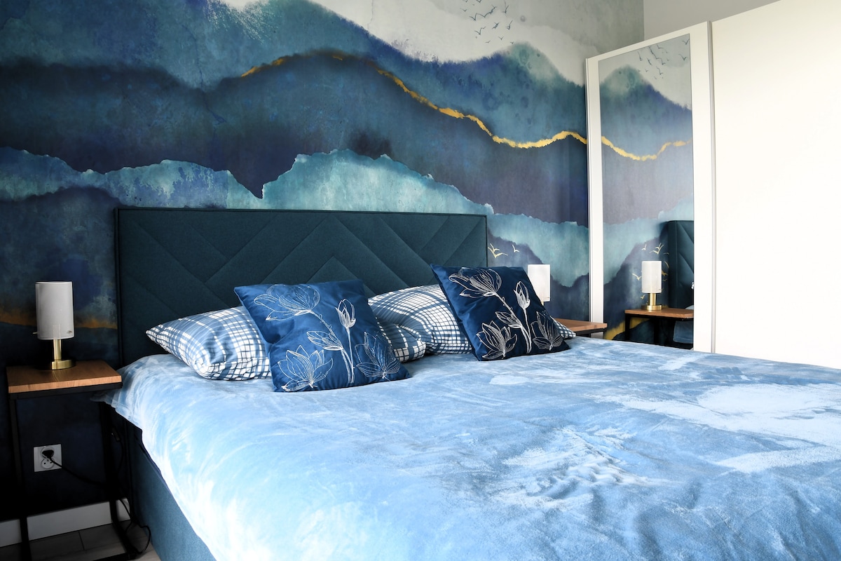 A spacious bed is dressed in a soft blue blanket, complemented by decorative pillows in contrasting designs. The wall behind features a watercolor mural with shades of blue and gold, while bedside tables are equipped with modern lamps. A full-length mirror reflects the room's aesthetic.