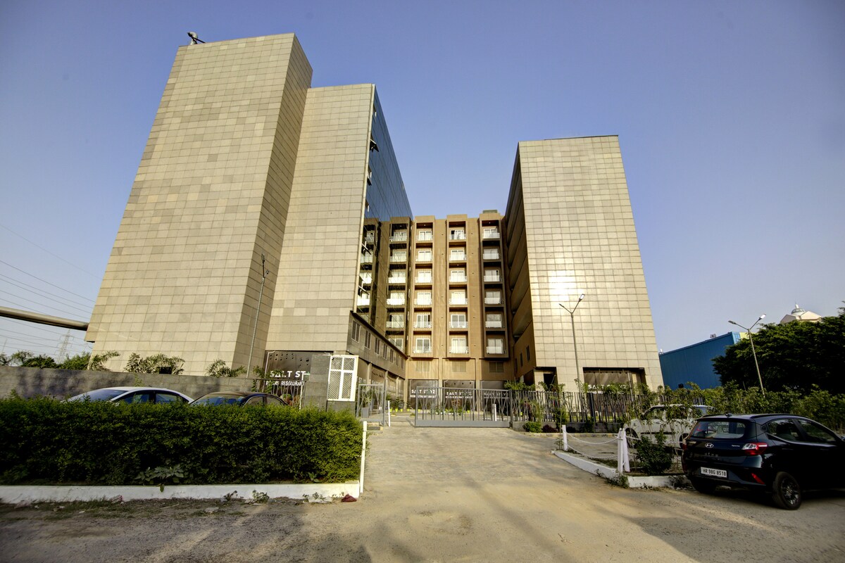 A modern building complex is depicted, characterized by large glass panels and a bronze façade. The structure includes multiple floors with well-defined windows. Surrounding greenery is visible, along with a paved pathway leading to the entrance. A clear blue sky enhances the overall appearance.