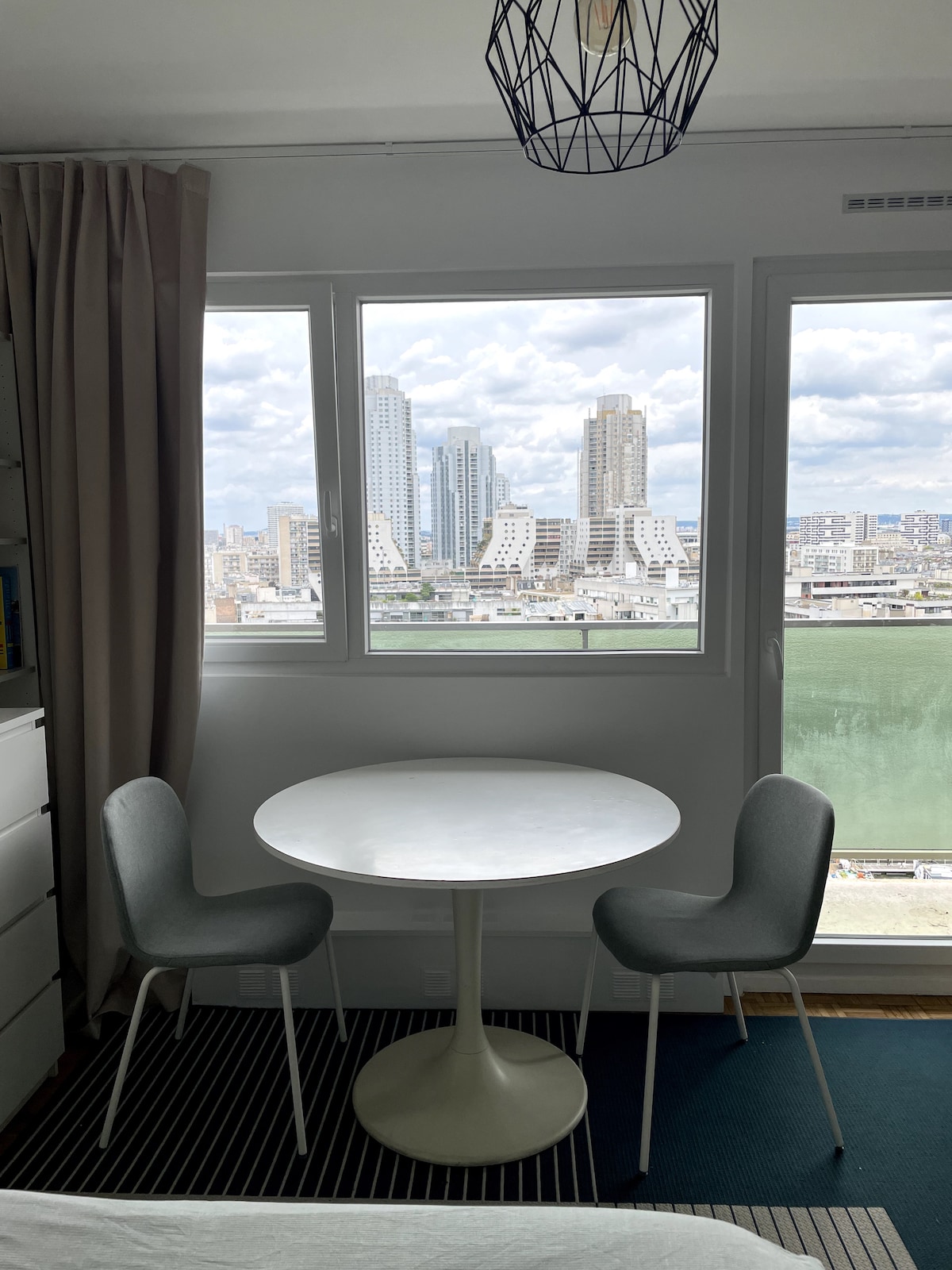 A round white dining table is positioned between two gray chairs, enhanced by large windows that offer a view of waterfront buildings and the city skyline. Natural light fills the space, highlighting the modern decor and inviting atmosphere.
